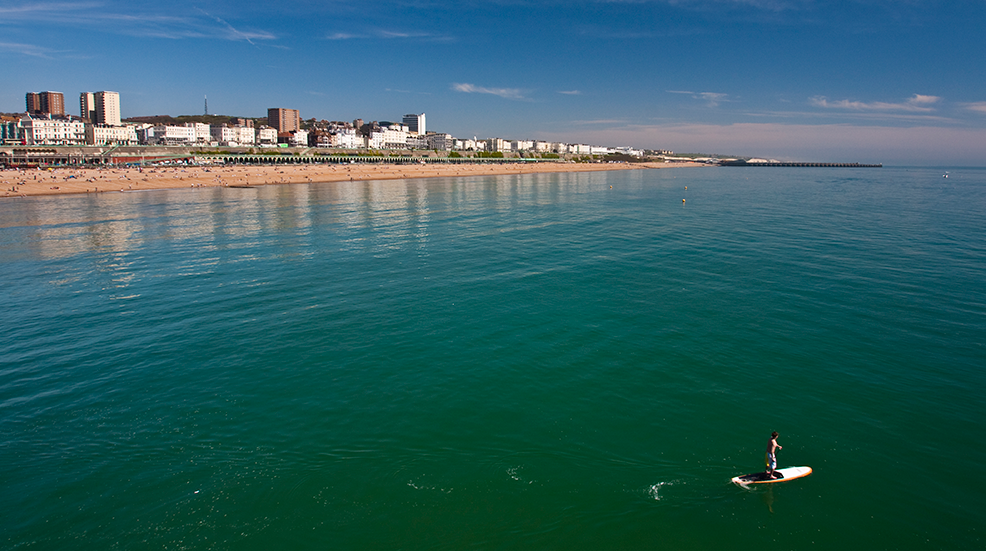 Birds eye view of Brighton seafront with the promenade reflecting in the water and a man stand up paddle boarding